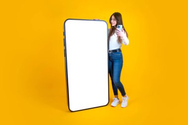Büyük akıllı telefon, uzun vücut boyunda beyaz kadın, akıllı telefona eğiliyor. Milenyum hanımefendisi elinde cep telefonu tutuyor ve büyük olanını boş ekranla maket için eğiyor. Sarı arkaplan, boşluğu kopyala. 