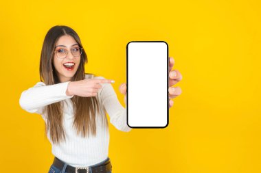 İşaret parmağı, güzel beyaz bir kızın portresi elindeki akıllı telefonu işaret ediyor. Boş beyaz ekran cep telefonu modeli. İzole edilmiş sarı arkaplan uygulamasına tepki gösteriliyor. 