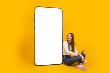 Uygulama tasarımı için bir model. Kafkas genç bayan boş ekranlı büyük bir cep telefonunun yanında oturuyor. Cep telefonu kullanıyor. Büyük ekranda görünüyor. İzole edilmiş sarı arkaplan, kopya tasarımı. 