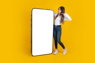 Kocaman boş ekranlı cep telefonunu işaret eden bir kadın. Tüm vücut uzunluğu, izole edilmiş sarı stüdyo arka planında duran genç bayanı hayrete düşürdü. Kot pantolon ve beyaz gömlek giyiyordu. Akıllı telefon modeli, boşluğu kopyala.