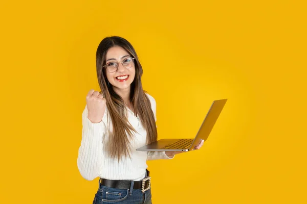 Woman holding laptop, celebrating with winner gesture. Beautiful young brunette office worker businesswoman 20s years old in basic white shirt and jeans standing over yellow background. Copy space.