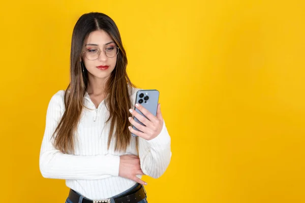 Serious woman, portrait of caucasian young serious woman. Businesswoman standing over isolated yellow background, copy space. Holding smartphone, looking screen. Thinking bad news, unhappy.