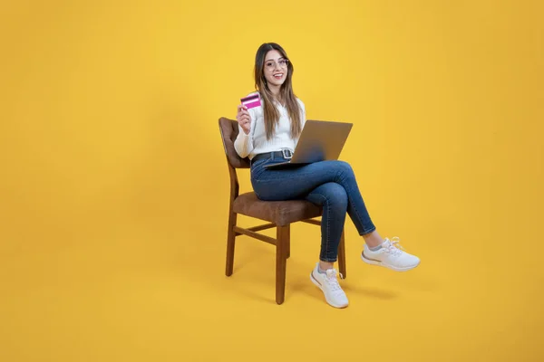 Holding credit card and laptop, full length body view of woman sitting on chair holding credit card and laptop. Buying goods via internet, online shopping concept image. Payment, remote banking idea.