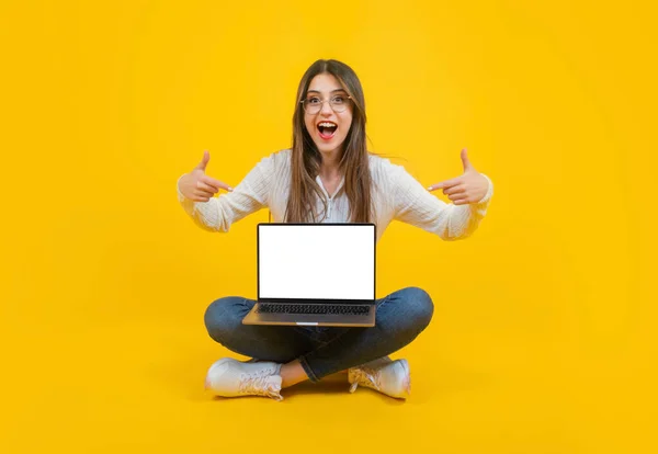 Laptop template, modern beautiful full length view woman sitting on ground. Cheerful brunette girl pointing fingers empty blank screen of notebook mockup. Smiling, showing portable pc. Copy space.