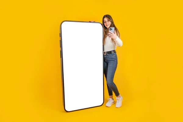 Blank screen big phone, full body young smiling woman holding smartphone and leaning blank screen big phone. Cellphone mockup. Female making video call. Wearing jeans Isolated yellow studio background