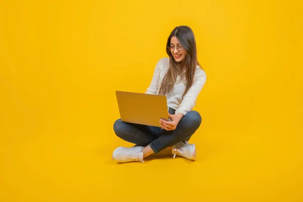 stock image Happy university student using her computer. Positive gesture, smiling looking her laptop screen. Office worker, full length body view caucasian girl sitting ground. Yellow studio background.