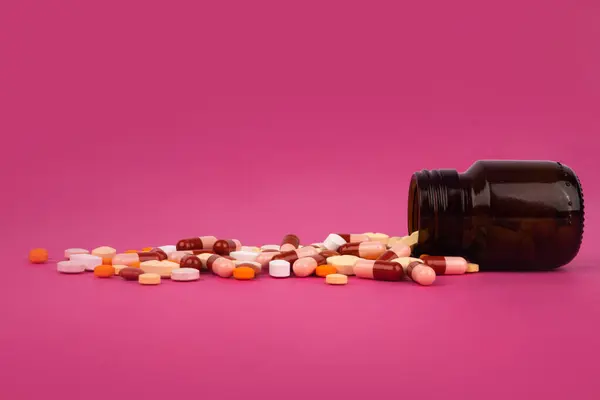 オレンジ色のガラス薬瓶からこぼれるカラフルな薬 サイドビュー画像 ピンクの背景に隔離された薬局の概念 コピースペース ビタミン鎮痛剤抗生物質治療サプリメント — ストック写真