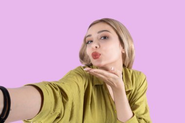 Hava öpücüğü yollamak, hava öpücüğü gönderen genç sarışın kızın yakın portresi. Yeşil tişörtlü güzel sarışın beyaz kadın izole açık mor arka plan pozu veriyor. Selfie çekimi.