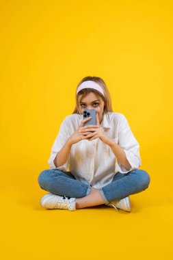 Akıllı telefon, tüm vücudu gören genç sarışın kız akıllı telefon kullanıyor. Zeminde çapraz bacaklı, sarı arka planda izole bir şekilde oturuyor, kopyalama alanı. Yüzünün yarısını cep telefonuyla kapat. İletişim.