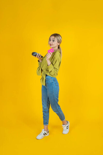 Holding wireless bank payment terminal, full body view blonde young girl holding wireless bank payment terminal and credit debit card. Isolated yellow background. Copy space. Cashless payment concept.