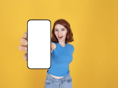Modern akıllı telefon, sevimli, kızıl saçlı, modern akıllı telefon gösteren beyaz bir kadın. Boş beyaz ekran cep telefonu modeli. İzole edilmiş sarı arkaplan, kopyalama alanı.