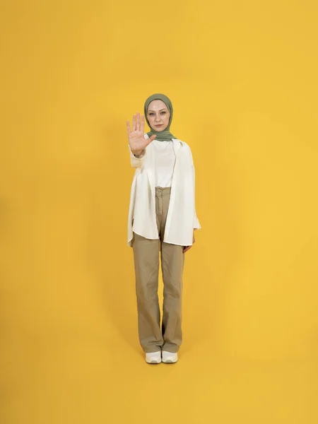 Muslim woman doing stop sign with palm of the hand. Freedom of religion, don\'t touch my head scarf. Warning expression negative, serious gesture. Full body front view girl standing yellow background.