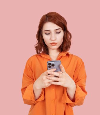 Akıllı telefon kullanan bir kadın, akıllı telefon kullanan genç ve güzel kızıl saçlı bir kadının portresini yakın çekim. Turuncu gömlek giy, pastel pembe arka plan stüdyo çekimlerinin üzerinde dur. Ciddi yüz ifadesi. 