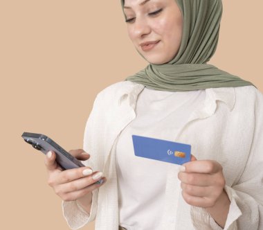 Kolay para transferi, tesettürlü genç müslüman kızın kolay para transferi portresi. Yüze odaklan. Elinde akıllı telefon ve kredi kartı tutan gülümseyen bir kadın. İnternetten alışveriş, vergi ödeme.
