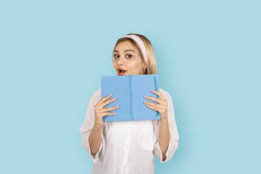 Çenesini kitapla kaplayan beyaz gömlekli genç sarışın kız, izole edilmiş mavi arka plan stüdyo portresi yapıyor. İnsanların yaşam tarzı mekanı taklit eder. Şaşırmış yüz ifadesi..