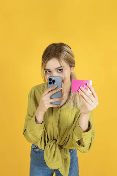 Using phone and credit card, portrait of young blonde teenage girl using phone and credit card. Positive expression, dressed casual, online shopping, e-commerce, isolated yellow background.