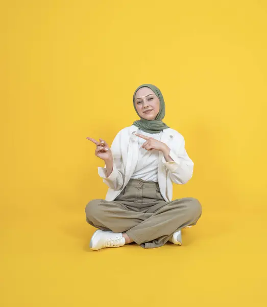 stock image Pointing showing copy space, young muslim caucasian woman in hijab head scarf sit floor legs crossed pointing showing copy space. Looking camera. Offer, recommend, advert, banner design concept image.