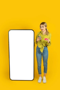 Tam vücut ön manzara genç sarışın kız büyük boş ekran cep telefonu modelinin yanında duruyor. Akıllı telefon ve kredi kartı tutuyor. Çevrimiçi alışveriş ödeme konsepti. Sarı arkaplan. 