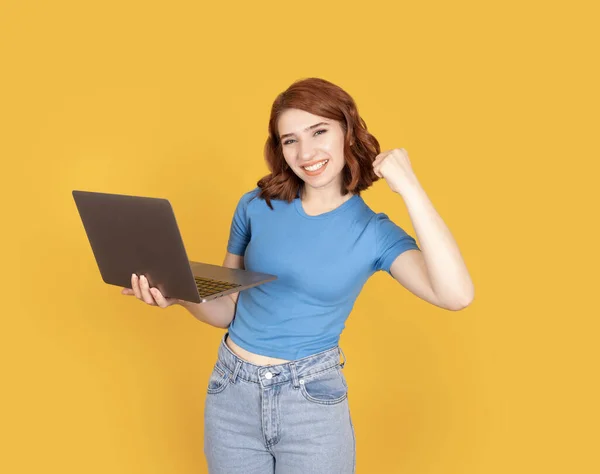 Young red bob hair business woman student smiling holding laptop pc computer. Freelance lady 20s blue t-shirt do winner gesture clench fist isolated yellow background studio portrait. Copy space.