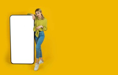 Yeni mobil uygulama öneriliyor. Beyaz tenli eğlenceli sarışın kız yeni mobil uygulama öneriyor. Genç bir kadın kocaman boş ekran akıllı telefonuna eğiliyor. Boşluğu kopyala, reklam tasarımı.