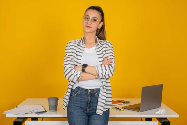 Kendine güvenen bir iş kadını, beyaz esmer, kendine güvenen, ofis masası kollarını katlamış bir iş kadını portresi. Gülümsüyor, menajer görüşmeye hazır. Bilgisayarla çalışıyorum. Sarı stüdyo arka planı.