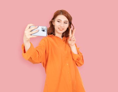 Barış işareti gösteren, güzel kızıl saçlı kadın gülümseyen ve barış işareti gösteren. Selfie çekmek, blog yazmak, sosyal medya. Elinde akıllı telefonuyla gülümseyen pozitif bir genç kız. İzole edilmiş pembe arkaplan.