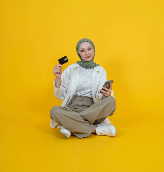 Hold plastic debit credit bank card,  full body view portrait of young caucasian muslim girl sit floor hold plastic debit credit bank card. Using mobile phone, smartphone for online shopping concept.