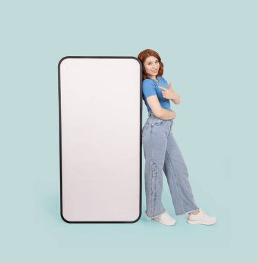 Kocaman boş ekranlı cep telefonu, tüm vücudu beyaz kadın kocaman boş ekranlı cep telefonunu işaret ederek ayakta duruyor. Modern akıllı telefon modeli. İzole edilmiş mavi arkaplan.