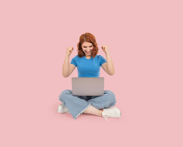 Woman celebrate clench fists, full body young caucasian sitting floor woman celebrate clench fists. Looking holding computer. Winner concept. Jubilant successful girl. Isolated over pink background.