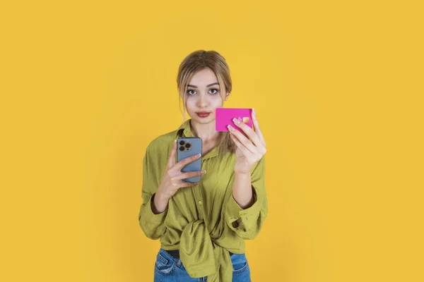 Woman hold credit bank card, young beautiful blonde woman hold credit bank card. Standing over yellow studio background. Hold mobile phone. 20s caucasian girl offer online shopping, payment concept.