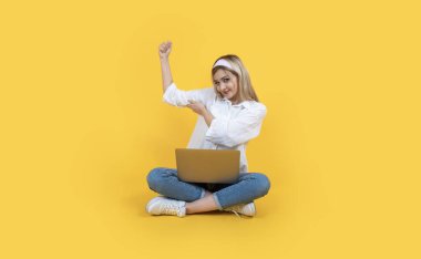 Elinizi sıkın, neşeli, mutlu, güzel, sarışın, sımsıkı sarılan kadın elinizi kaldırın. Dizüstü bilgisayar, zemin bacak bacak üstüne at. Kazanan konsept, mutluluk heyecanını ifade eder. Sarı arkaplan.
