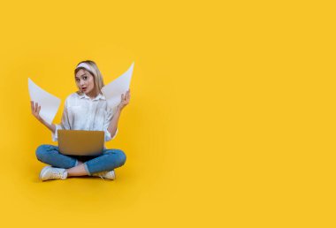 Elinde dizüstü bilgisayarı olan bir kadın, tüm vücudu gören genç beyaz bir kadın elinde dizüstü bilgisayarla belgeleri tutuyor. Sarı stüdyo arkaplanının üzerine otur, kopyalama alanı. Kameraya bak, şaşkın, şaşkın.. 