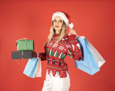 Sunum konsepti satın almak, hediye alan çekici genç bir kadının portresi. Kazak ve Noel Baba 'nın şapkasını giy. Kırmızı stüdyo geçmişi. Hediye kutuları ve alışveriş torbaları. Noel fikri.