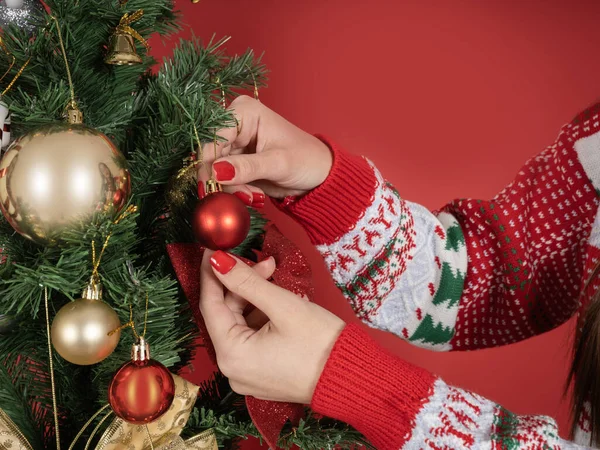 Decorating Christmas tree, close up image of woman hands decorating  Christmas tree. Holding red ball. Wear knitted sweater. Standing over red studio background. Happy new year holiday concept idea.