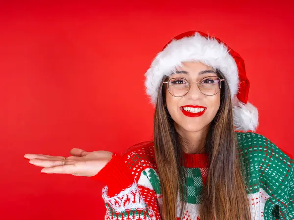 Woman showing open palm, portrait of happy young caucasian woman showing open palm. Wear santa hat, Christmas knitted sweater standing over isolated red studio background. Holding invisible product.