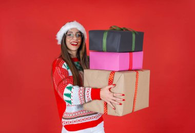 Noel hediye kutuları, örgü kazaklı beyaz kadın ve Noel Baba şapkası içinde bir sürü hediye kutusu. Kış tatili, mutlu yıllar konsepti. İzole kırmızı stüdyo arka planı. Boşluğu kopyala.