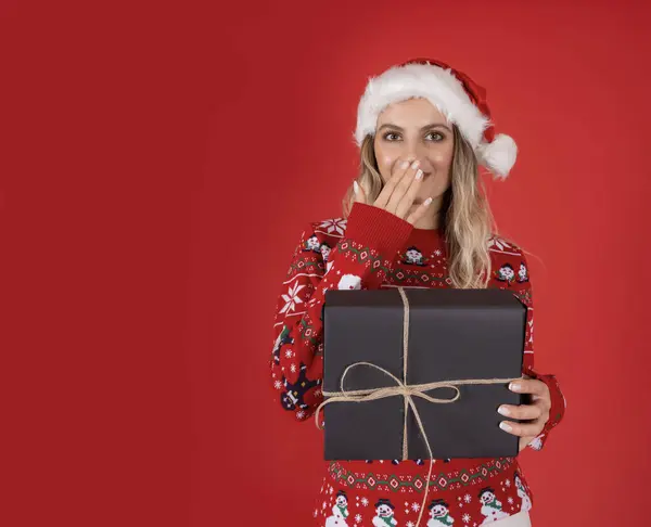 Happy shocked woman,  portrait of young caucasian happy shocked woman holding present gift box wear knitted Christmas sweater and Santa\'s hat standing over isolated red studio background. Copy space.