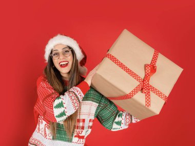 Kadının elinde büyük bir hediye kutusu, genç kadının portresi büyük bir hediye kutusu var. Noel süveteri, Noel Baba şapkası giy. Heyecanlı, gülümseyen, meraklı kız. Mutlu yeni yıl kış tatili konsepti.