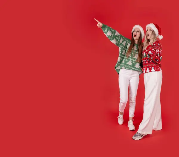 Excited two woman, full body studio portrait of excited two woman. Pointing copy space, isolated red studio background. Recommend, offer, Christmas advertising concept image.