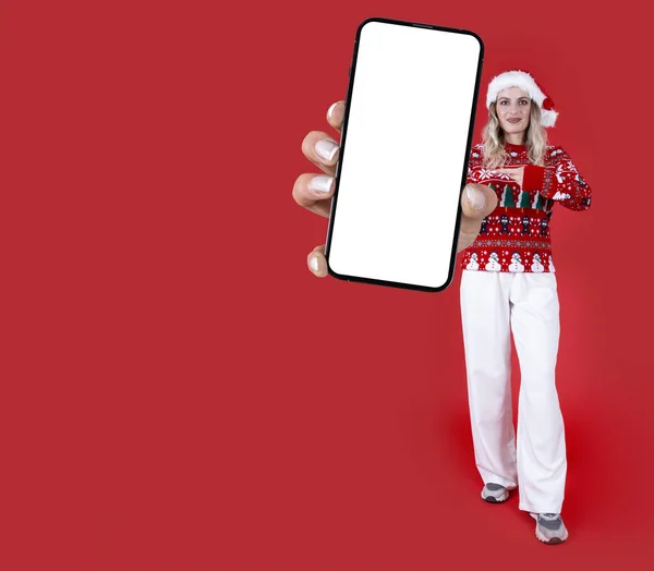 Merry young full body woman in knitted Christmas sweater and Santa hat hold show use big mobile phone mock up blank screen isolated red studio background. happy new year winter holiday concept idea.
