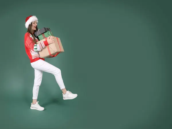 Hold present box, full body overjoyed young woman hold present box. What Santa hat, knitted Christmas sweater. Happy new year celebration holiday concept. Isolated green studio background. Copy space.