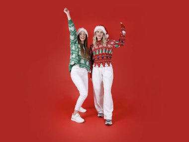 İki kadın birlikte dans ediyor, tüm vücudu eğlenceli iki beyaz kadın birlikte dans ediyor. Noel Baba şapkası tak, Noel süveteri giy. Ellerini aç, hava al. Mutlu yıllar, kırmızı arka plan. Boşluğu kopyala.