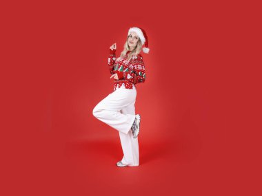 Çekici Noel Baba kızı, genç sarışın, Avrupalı çekici Noel Baba kızı portresi. Noel kazağı, şapka ve beyaz pantolon giy. Hareket et, yürü, dans et. İzole kırmızı stüdyo arka planı.