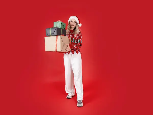 Holding stack of gift boxes, full body studio portrait of young Santa woman holding stack of gift boxes. Celebrating Christmas winter holiday. Shopping taking giving gift. Isolated red background.