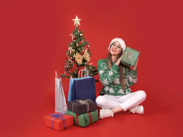 Stock image Shaking gift box, full body view young Santa woman sit near decorated Christmas tree shaking gift box. Wondering what's in inside present. Winter holiday, happy new year celebration concept idea.