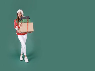 Elinde bir yığın hediye kutusu, tüm vücudu heyecanlı beyaz bir kadın elinde bir yığın hediye kutusu tutuyor. Noel Baba şapkası, örgü Noel süveteri, beyaz pantolon giy. Tatili kutluyoruz. Boşluğu kopyala.