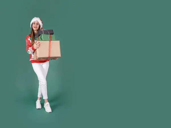 Holding stack of gift boxes, full body excited pretty caucasian attractive woman holding stack of gift boxes. Wear Santa hat, knitted Christmas sweater, white pants. Celebrating holiday. Copy space.