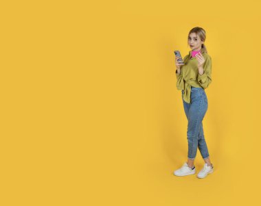 Cep telefonu ve kredi kartı tutuyor. Tam vücut görüşlü, sarı arka planda duran beyaz tenli sarışın kadın. İnternetten alışveriş, ödeme fikri. Boşluğu kopyala.