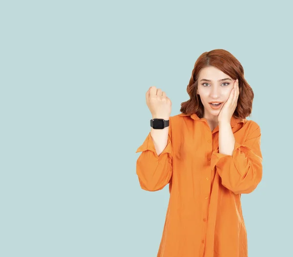 Showing smart watch screen, young shocked excited woman showing smart watch screen. Portrait of red bob hair girl touching her cheek, standing light blue background, copy space. Technology concept.
