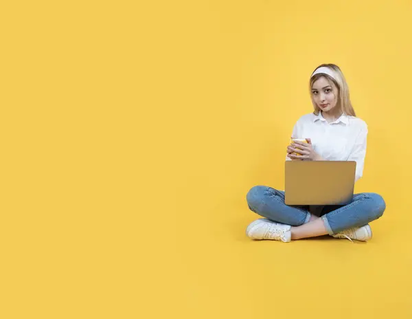 Young woman sit floor hold use laptop drinking coffee. Full length body view caucasian blonde girl student sit legs crossed. Isolated yellow background. Copy space. Education concept idea.